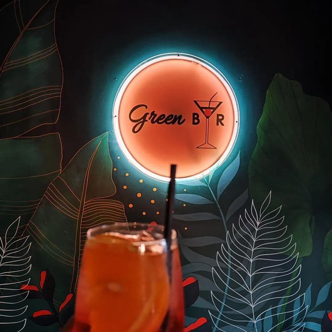Green Bar neona zīme ar kokteiļa glāzi uz tumši zaļas sienas ar tropu lapām, priekšplānā dzēriens ar salmiņu